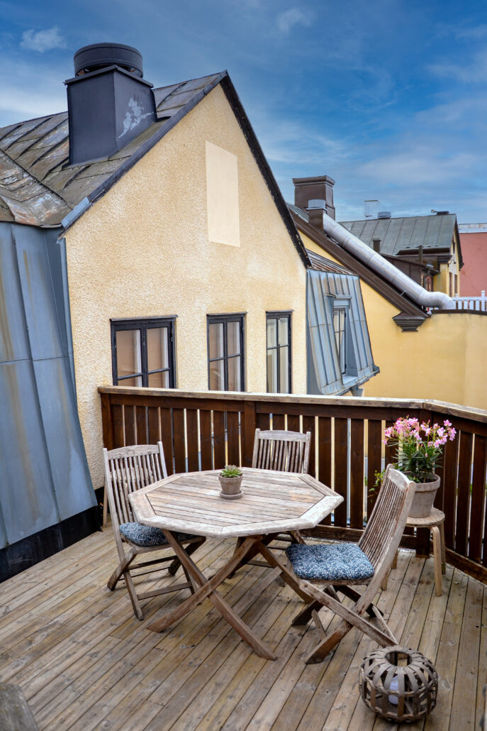 Takterassen med plats för 6 personer vätter mot innergården och parkeringen. Bild på Terassen med ett borden och tre stolar till, sittgrupp i trä. Dekorerad med blommor. Sittkuddar på stolarna. Blå himmel och gula hus och takåsar i bakgrunden. Träterass.